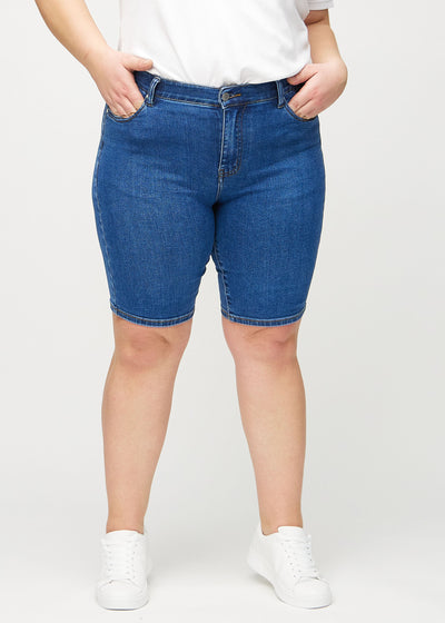 Mørk denim skinny middle shorts, modelnavn Oceans, som sidder tæt til låret og slutter lige over knæet på en plus-size model, set forfra.